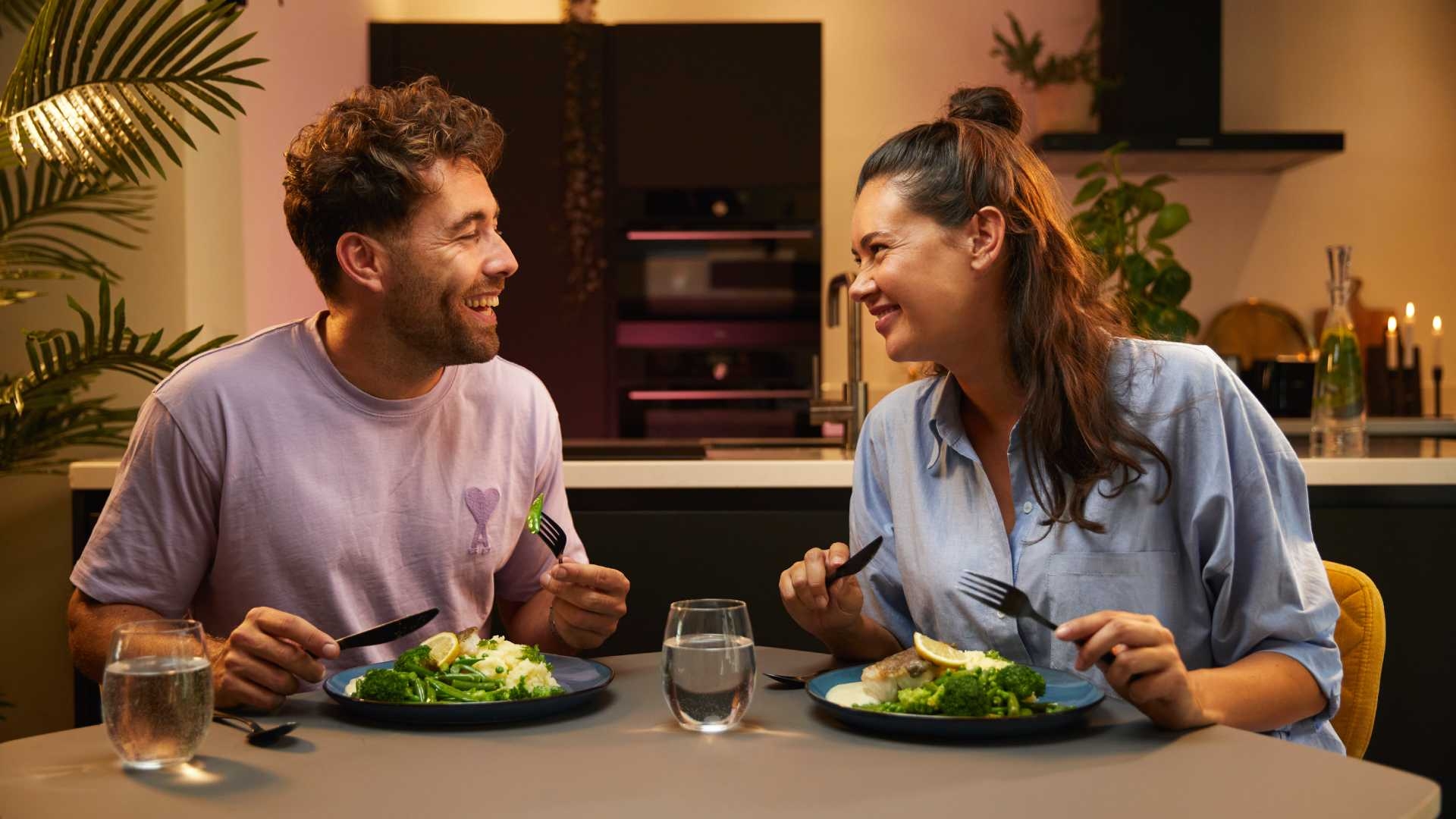 Een stel dat samen aan tafel eet en elkaar lachend aankijkt, met een keuken op de achtergrond